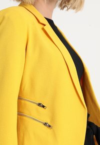 Žlutý blazer s hladkou texturou, s upraveným střihem, klopami a stříbrnými kapsami na zip po stranách.