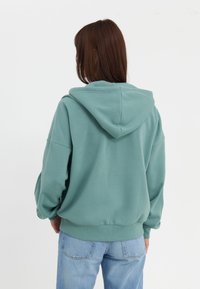 Donna con capelli castani lisci che indossa un ampio hoodie teal e jeans di un blu chiaro, rivolta di spalle su uno sfondo bianco semplice.
