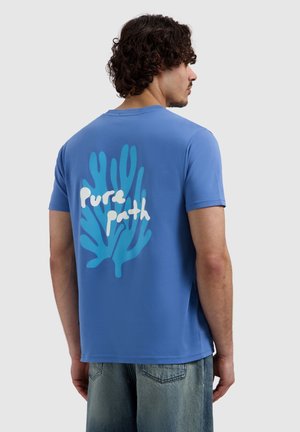 Man met krullend donker haar die een blauw T-shirt draagt met een grote blauwe koraalafbeelding en witte tekst "Pure Path" op de rug, gecombineerd met vervaagde spijkerbroek.