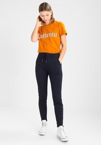 Oransje bomull t-skjorte med "Authentic" tekst, kombinert med marineblå joggebukser med elastisk midje og lommer, brukt med hvite sneakers.