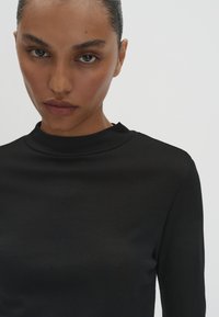 Frau mit dunkler Haut und zurückgegelltem Haar, die ein schwarzes Langarmoberteil mit hohem, rundem Ausschnitt vor einem einfarbigen Hintergrund trägt.