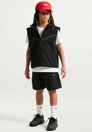 Niño con cabello largo que lleva una gorra roja, chaleco sin mangas negro, camiseta blanca, pantalones cortos negros, calcetines blancos y zapatillas negras, de pie.