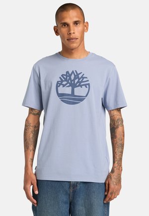 Uomo con tatuaggi che indossa una maglietta azzurro chiaro con un logo di un albero blu scuro e jeans blu, in piedi davanti a uno sfondo semplice.