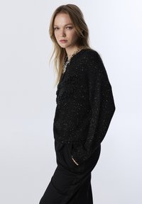 Černý pletený svetr s texturovanými, třpytivými nitěmi. Má uvolněný střih a výrazný třásňový detail na přední straně. Nosit s černými kalhotami.