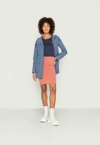 Mulher a usar uma jaqueta azul, blusa azul-marinho, saia cor-de-pêssego e botas brancas, de pé com uma mão no bolso da saia, contra um fundo simples.