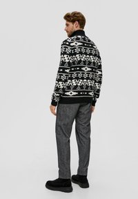 Schwarzer und weißer gemusterter Pullover mit geometrischen Designs, kombiniert mit grauen strukturierten Hosen und schwarzen Wildlederstiefeln.