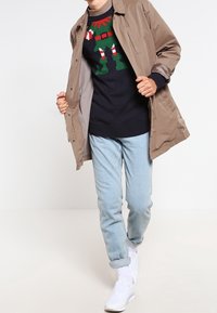 Veste légère beige, pull bleu marine avec motif vert et rouge, jean bleu clair, et baskets blanches. Tenue décontractée avec des textures superposées.