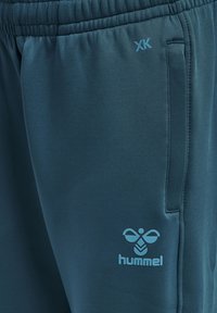 Teal Hummel sweatpants med elastisk midja, sidoficka och en ljusblå logotyp som har en symmetrisk design och "XK"-detalj.