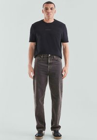 Mann, der mit dem Gesicht nach vorne steht, trägt ein schwarzes T-Shirt mit kleinem Text und ausgewaschenen schwarzen Jeans sowie schwarze Sneakers vor einem schlichten hellen Hintergrund.