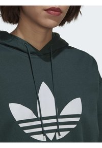 Grön hoodie med en stor vit Adidas trefoil-logotyp och tre horisontella ränder. Justerbara dragkedjor vid halsen. Mjuk tygkvalitet.