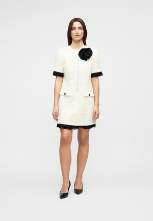 Femme de face portant une robe blanche texturée avec une bordure à volants noire et un grand détail fleur noire sur la poitrine, talons noirs.