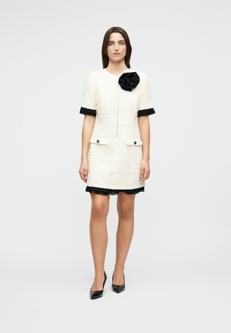 Γυναίκα στέκεται αντικρίζοντας μπροστά, φορώντας ένα λευκό φόρεμα με υφή και μαύρη φραμπαλά περιμετρικά, με μια μεγάλη μαύρη λουλουδένια λεπτομέρεια στο στήθος και μαύρες γόβες.