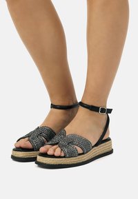 Sandali wedge neri con texture intrecciata, cinturino alla caviglia e suola in giuta intrecciato. Caratterizzati da un design con fasce incrociate e dettagli lucidi sulla parte superiore.