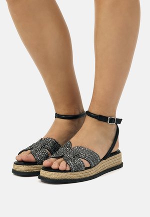 Sandali wedge neri con texture intrecciata, cinturino alla caviglia e suola in giuta intrecciato. Caratterizzati da un design con fasce incrociate e dettagli lucidi sulla parte superiore.