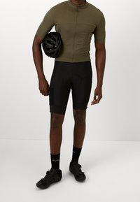 Maillot de cyclisme olive à manches courtes avec une fermeture éclair, associé à un short rembourré noir. Des chaussures de cyclisme noires et des chaussettes complètent la tenue. La personne tient un casque noir.