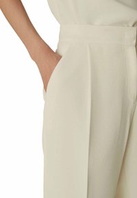 Pantaloni color crema realizzati in tessuto leggero con una consistenza liscia. Presentano una tasca laterale e una vestibilità a vita alta sartoriale con pinces.