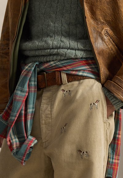 Giacca di pelle marrone sopra un maglione verde lavorato a trecce, camicia a quadri annodata in vita, pantaloni in velluto beige con dettagli ricamati a forma di cane.