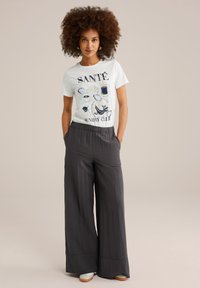 White t-shirt with graphic print featuring food items and text. Loose-fitting grey wide-leg pants. Model stands with hands in pockets.