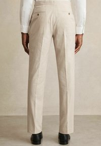 Beige, texturált nadrág függőleges csíkokkal, szabott fazonnal, két gombos hátsó zsebbel és fekete cipővel.
