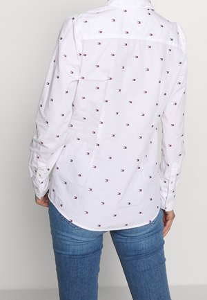 Weißes Langarmhemd mit kleinen roten und marineblauen Logo-Mustern, getragen mit blauen Jeans, von hinten gesehen.