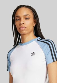 Adidas majica u bijeloj boji s svijetlo plavim rukavima, koja ima crne trostruke točke i mali crni logo na prsima.