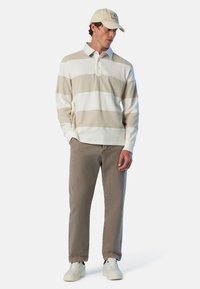 Mann in beigem und weißen gestreiften Langarm-Polo, taupefarbenen Hosen, weißen Sneakers und beigem Hut, steht mit den Händen in den Taschen und schaut nach unten.