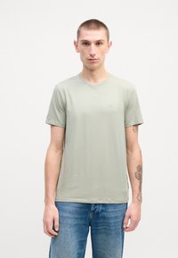 T-shirt vert clair en tissu doux et lisse avec un col rond et des manches courtes. Présente un petit logo près de la poitrine. Porté avec un jean bleu.