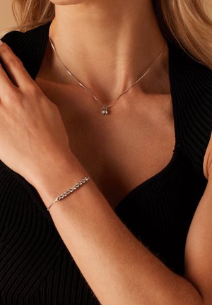 Zilveren halsketting met een kleine hanger en een bijpassende zilveren armband met meerdere ronde accenten, gedragen op een bloot arm tegen een zwarte achtergrond.