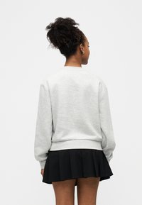 Femme de dos avec des cheveux bouclés en chignon, portant un sweat-shirt gris clair et une jupe plissée noire courte sur un fond blanc uni.