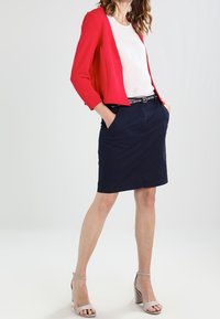 Červený zkrácený blazer přes bílý top, doplněný sukní po kolena v námořnické modři. Šedé sandály na vysokém podpatku dokončují outfit.