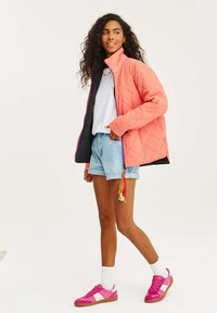 Prošívaná růžová bunda s černým podšívkou a kontrastním zipem, spárovaná se světlými modrými šortkami a růžovými teniskami s bílým akcentem.