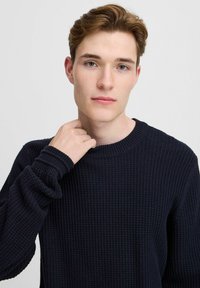 Pulover tricotat de culoare bleumarin, cu o suprafață texturată, guler rotund și manșete cu riduri. Purtați de o persoană care își ajustează gulerul.