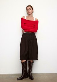 Roter, gerippter Off-Shoulder-Pullover kombiniert mit braunen, plissierten Shorts und braunen Cowboystiefeln mit geprägten Details. Schlichter Hintergrund.
