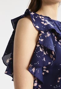 Marinblus med blommigt mönster som har volangärmar, tillverkad av slätt tyg med rosa blomstertryck och en figurnära passform.