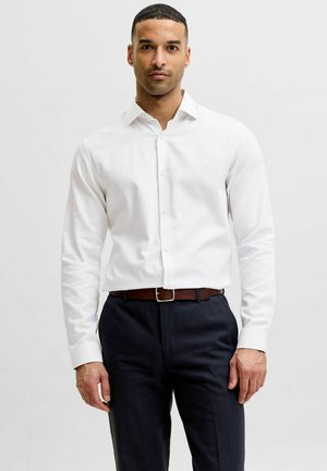 Homme aux cheveux courts portant une chemise blanche à manches longues boutonnée, un pantalon sombre et une ceinture marron, debout devant un fond uni.