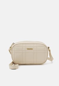 Béžová prošívaná crossbody taška se zlatým logem, vybavená zipovým zavíráním a odnímatelným popruhem pro různé způsoby nošení.