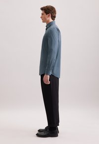 Camicia a maniche lunghe blu con motivo, abbinata a pantaloni e scarpe neri, che mostrano un taglio moderno e una texture liscia. Vista di profilo.