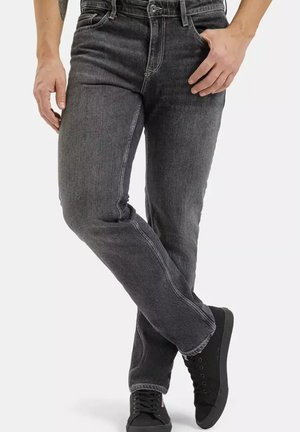 Mężczyzna w wyblakłych czarnych dżinsach i czarnych trampkach, stojący z skrzyżowanymi nogami i jedną ręką spoczywającą na biodrze.