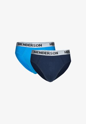 Oberkörperfreier Mann mit kurzen dunklen Haaren, der marineblaue Slips mit weißem Bund trägt, auf dem "HENDERSON" steht, steht mit den Händen hinter dem Rücken.