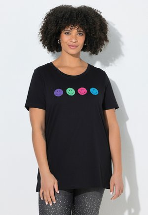 Kvinde med krøllet hår iført en sort t-shirt med fire farverige smiley-ansigter og mørke bukser med hvide prikker, stående mod en lys baggrund.