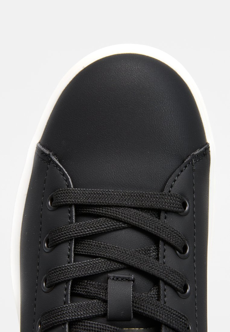 Zapatillas sintéticas negras con punta redonda, cordones negros y superficie texturizada. Suela de goma blanca con costuras detalladas cerca de los ojales.