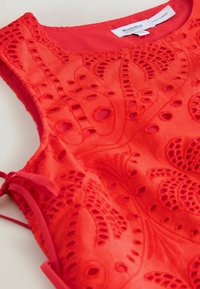 Top rouge sans manches avec broderie florale découpée complexe et détail nœud sur le côté, en tissu léger.
