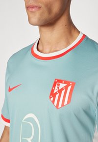 Camiseta deportiva azul claro con un cuello redondo rojo y blanco, que presenta un emblema rojo con estrellas y rayas, y un logo de Nike en rojo.