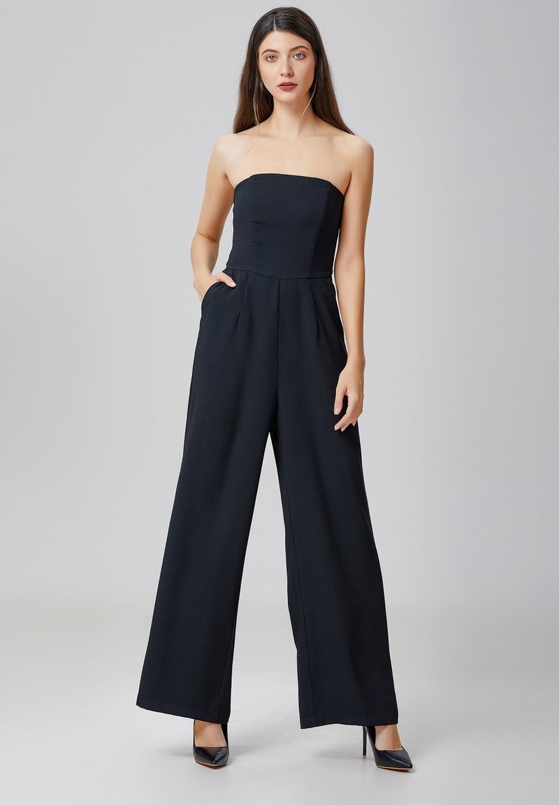 Combinaison noire sans bretelles avec un corsage ajusté, des jambes larges, des poches latérales et une texture lisse. Accompagnée de talons hauts noirs.