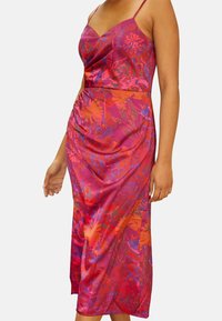 Nainen, joka pukeutunut hihattomaan, vaaleanpunaiseen ja oranssiin kukkakuosiseen midimekkoon, jossa on ohuet hihnat, yksinkertaisen valkoisen taustan edessä.