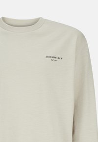 Ljust beige sweatshirt med rund halsringning, med en liten svart logotyptext som läser "JJ DENIM CREW Est. 1990" på vänster bröstområde.