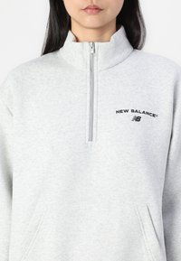 Lys grå hettegenser med halv-zip krage, med en frontlomme og svart "New Balance" tekstlogo. Myk fleece tekstur, løs passform.