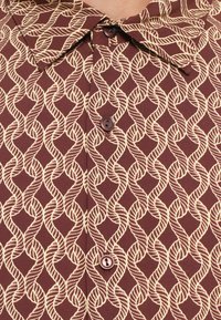 Pruun särk kreemja nöörimustriga, millel on terav krae ja nööbid. Materjal on sileda tekstuuriga.