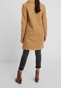 Donna in piedi di spalle, che indossa un cappotto camel in tessuto teddy, jeans neri arrotolati e stivali alla caviglia in pelle di serpente con tacco a blocco.