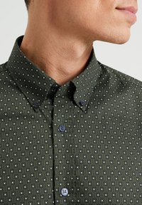 Zielona koszula w wzory z usztywnionym kołnierzykiem, z geometrycznym nadrukiem w białym i niebieskim kolorze. Z przodu widoczne detale na guziki.